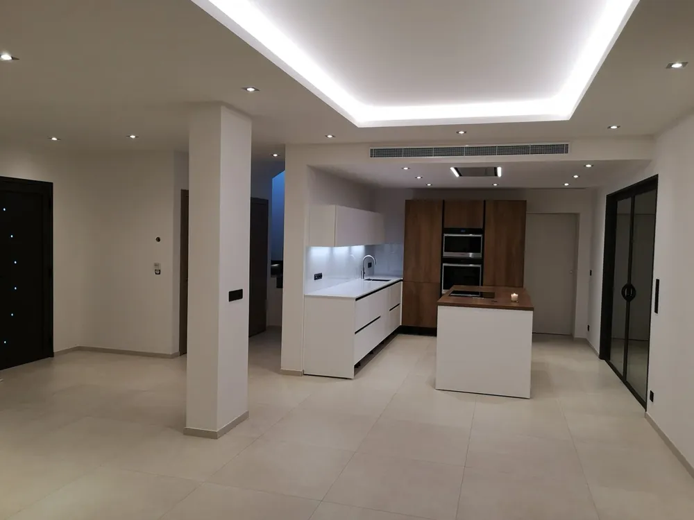 Cuisine ouverte rénovée avec îlot central, faux plafond à éclairage indirect et grand carrelage clair.