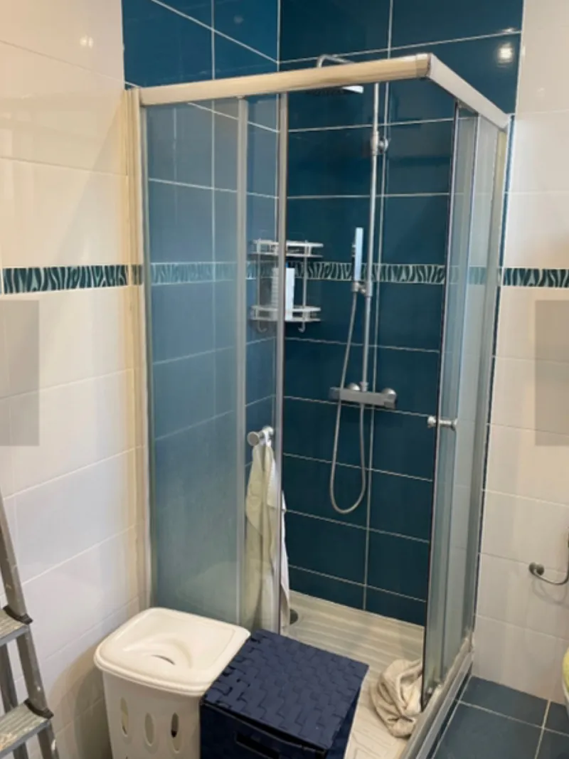 Salle de bain avant travaux : faïence bleue, cabine de douche d'angle et sol carrelé sombre.